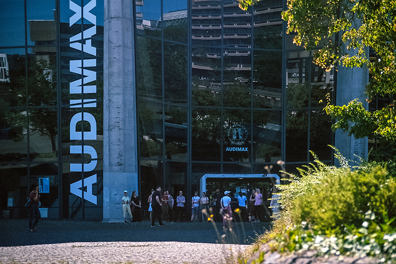 Foto: Eine Gruppe von Personen steht vor einem spiegelnden Gebäude. Von unten nach oben ist der weiße große Schriftzu "Audimax zu lesen". Am rechten Rand des Fotos befinden sich grüne Pflanzen und Bäume.