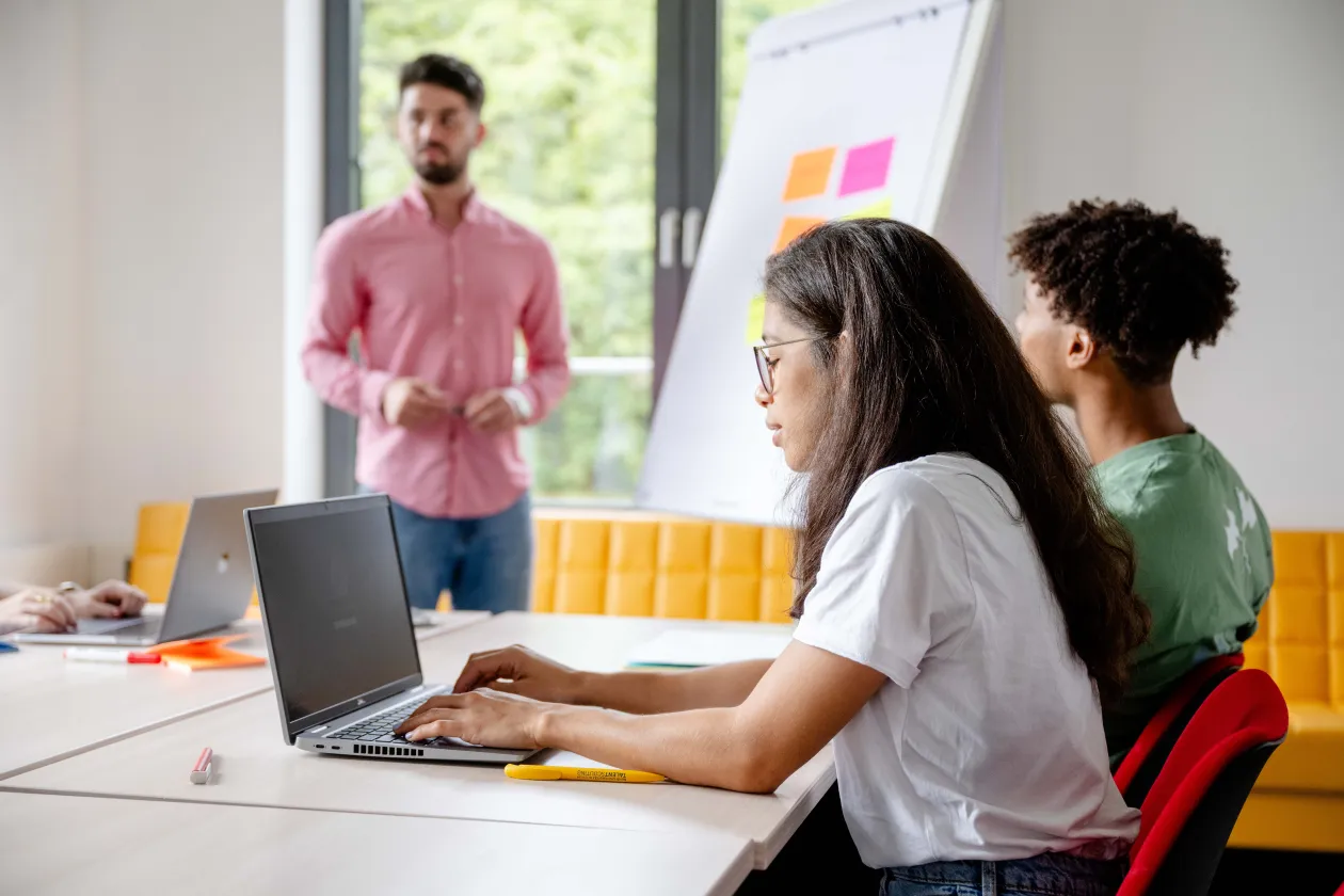 In einem Seminarraum steht der Dozent vorne neben einer Flipchart und die Studierenden sitzen an Tischen mit Heften und Notebooks.