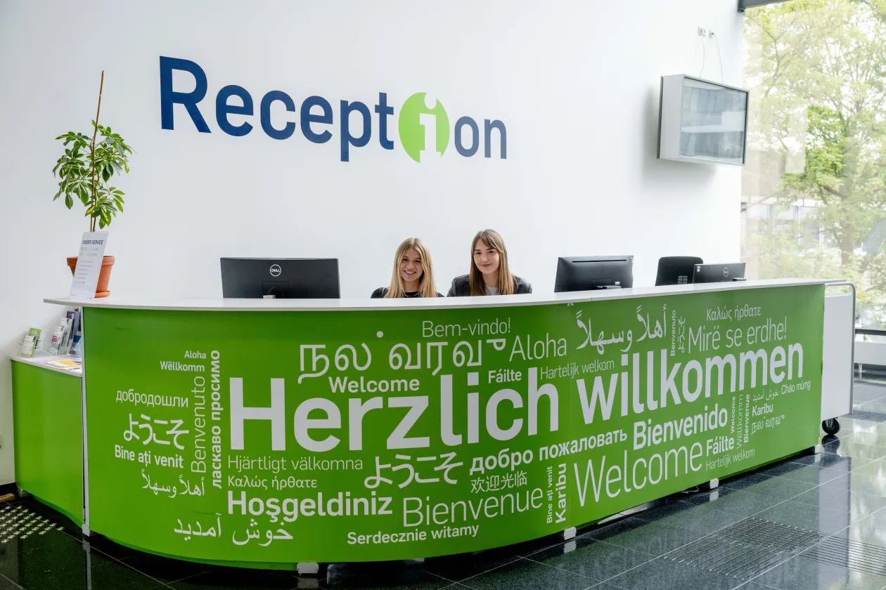 Foto: Hinter einer grünen Theke mit dem weißen Schritfzug "Herzlich willkommen" in vielen unterschiedlichen Sprachen und Schriften sind zwei sitzende Personen und drei Monitore zu sehen. Auf dem linken Ende der Theke steht eine Pflanze. An der Wand steht der Schriftzug "Reception" und es hängt ein Monitor an der Wand.