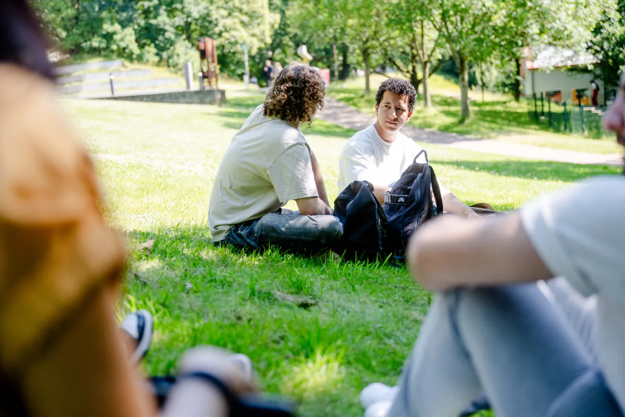 Foto: Vier Personen sitzen auf einer Wiese. Von zwei Personen sieht man nur die Schultern, die sich einmal links und einmal rechts im Foto befinden. Man schaut zwischen ihren Schultern durch auf zwei weitere Personen, die gut sichtbar nebeneinander auf der Wiese sitzen, sich anschauen und miteinander sprechen.
