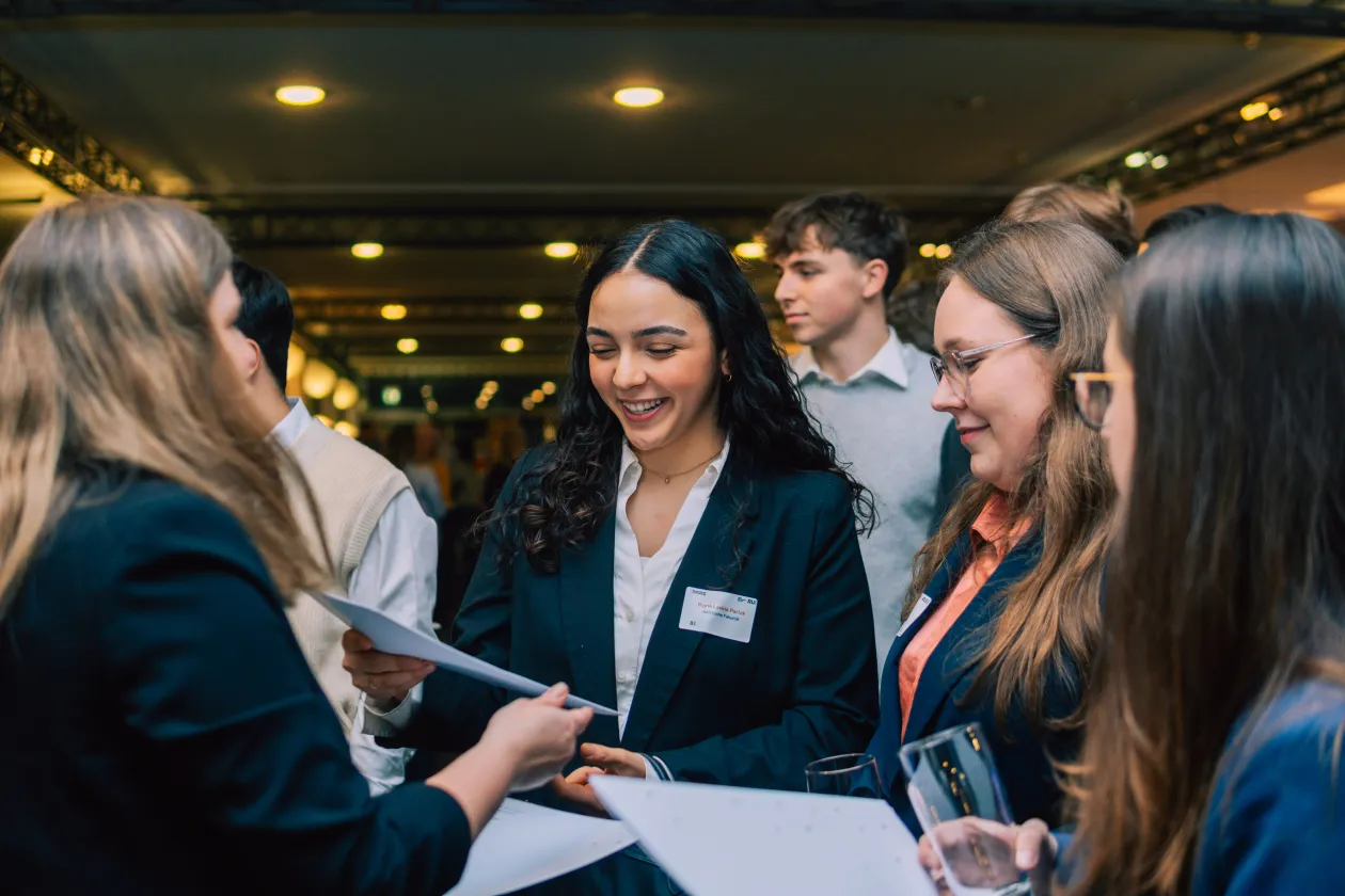 Foto: Eine Frau überreicht einer anderen Frau ein Dokument. Sie lächelt. Daneben stehen weitere Personen.