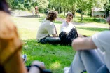 Foto: Vier Personen sitzen auf einer Wiese. Von zwei Personen sieht man nur die Schultern, die sich einmal links und einmal rechts im Foto befinden. Man schaut zwischen ihren Schultern durch auf zwei weitere Personen, die gut sichtbar nebeneinander auf der Wiese sitzen, sich anschauen und miteinander sprechen.