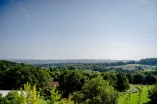 Foto: Blick in ein grünes Tal mit Bäumen bei Sonnenschein, es ist unter Bäumen ein Haus mit rotem Dach zu sehen.