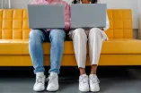 Foto: Zwei Personen sitzen nebeneinander. Beide haben einen aufgeklappten Laptop auf dem Schoß. Man sieht nur Füße und Beine der Personen sowie die Rückseite der Laptops.