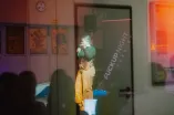 Foto durch eine Glasscheibe hindurch. Zu sehen ist eine Frau, die etwas auf einer Bühne erzählt.