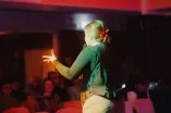 Rückansicht einer weiblich gelesenen Person, die auf einer Bühne steht und mit einem Mikrofon in der Hand Richtung Publikum spricht