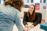Foto: Eine Person sitzt hinter einer Theke, man sieht sie von vorne. Eine andere Person steht vor der Theke, man sieht ihren Rücken. Beide Personen zeigen auf eine Karte des RUB-Campus, die auf der Theke liegt. Die Person hinter der Theke schaut die andere Person lächelnd an.