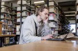 Junge männlich gelesene Person sitzt mit traurigem Gesicht vor einem aufgeklapptem Laptop. Im Hintergrund sind Bücherregale zu sehen.