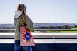 Das Foto zeigt eine Person auf der Mensaterrasse in der Rückenansicht, von der Schulter hängt ein RUB-Beutel mit Audimax-Motiv im 60er Jahre-Stil