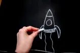 Das Foto zeigt eine Hand, die mit Kreide eine Rakete an eine Tafel malt.