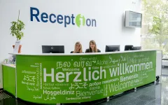 Foto: Hinter einer grünen Theke mit dem weißen Schritfzug "Herzlich willkommen" in vielen unterschiedlichen Sprachen und Schriften sind zwei sitzende Personen und drei Monitore zu sehen. Auf dem linken Ende der Theke steht eine Pflanze. An der Wand steht der Schriftzug "Reception" und es hängt ein Monitor an der Wand.