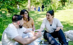 Das Foto zeigt zwei junge Frauen und einen jungen Mann die zusammen auf einer Decke auf einer Wiese sitzen und zusammen reden und lachen.