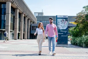 Foto: Eine Frau und ein Mann gehen auf dem betonierten Campus der Ruhr-Universität nebeneinander her. Sie sind frontal zu sehen, sie unterhalten sich und lachen. Es ist sonnig. Im Hintergrund sieht man ein Schild mit der Aufschrift "Willkommen" in mehreren Sprachen.
