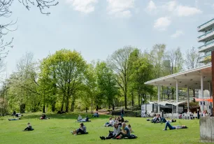 Foto: Studierende sitzen auf einer grünen Wiese im Sonnenschein. Rechts steht ein Gebäude mit Sitzplätzen davor.