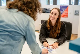 Foto: Eine Person sitzt hinter einer Theke, man sieht sie von vorne. Eine andere Person steht vor der Theke, man sieht ihren Rücken. Beide Personen zeigen auf eine Karte des RUB-Campus, die auf der Theke liegt. Die Person hinter der Theke schaut die andere Person lächelnd an.
