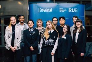 Foto: Mehrere Personen stehen lächelnd vor einem blauen Aufsteller. Auf dem Aufsteller befinden sich die Logos vom Deutschlandstipendium und von der Ruhr-Universität Bochum.