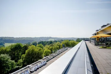Foto: Terrasse mit Sitzplätzen, Schirmen und einem Geländer, daneben Blick in ein Tal mit vielen grünen Bäumen, es ist sonnig.