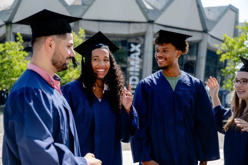 Mehrere Personen mit Abolsventenhüten stehen vor dem Audimax der Ruhr-Universität Bochum und unterhalten sich.