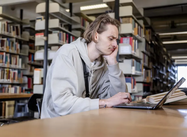 Junge männlich gelesene Person sitzt mit traurigem Gesicht vor einem aufgeklapptem Laptop. Im Hintergrund sind Bücherregale zu sehen.