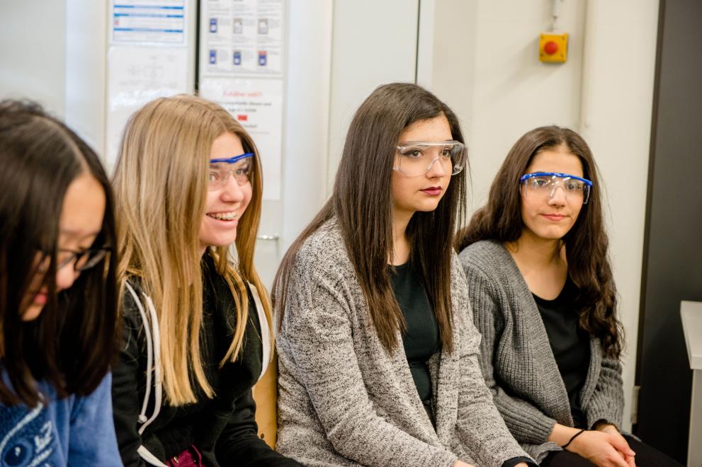 Vier Schülerinnen sitzen in einer Reihe und tragen Schutzbrillen.