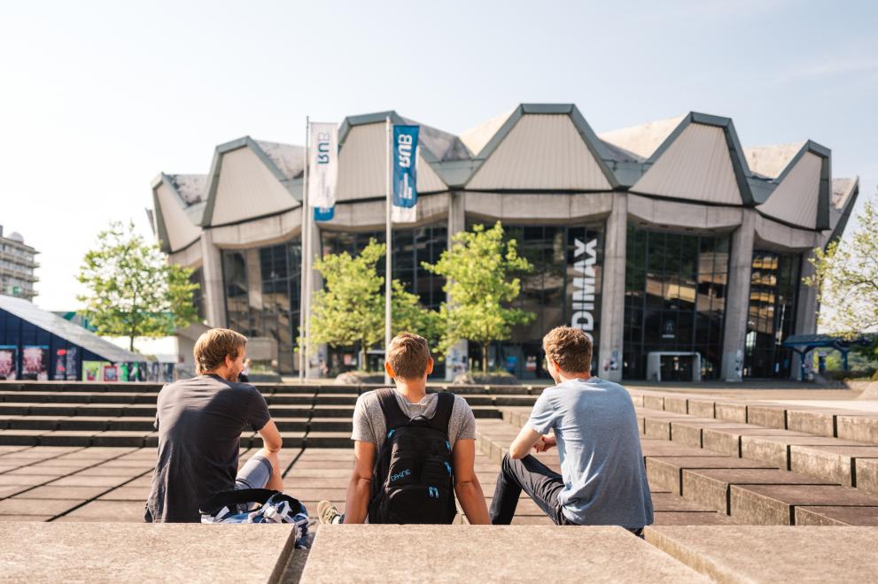 Drei Studierende sitzen vor dem Audimax der Ruhr-Unviersität.