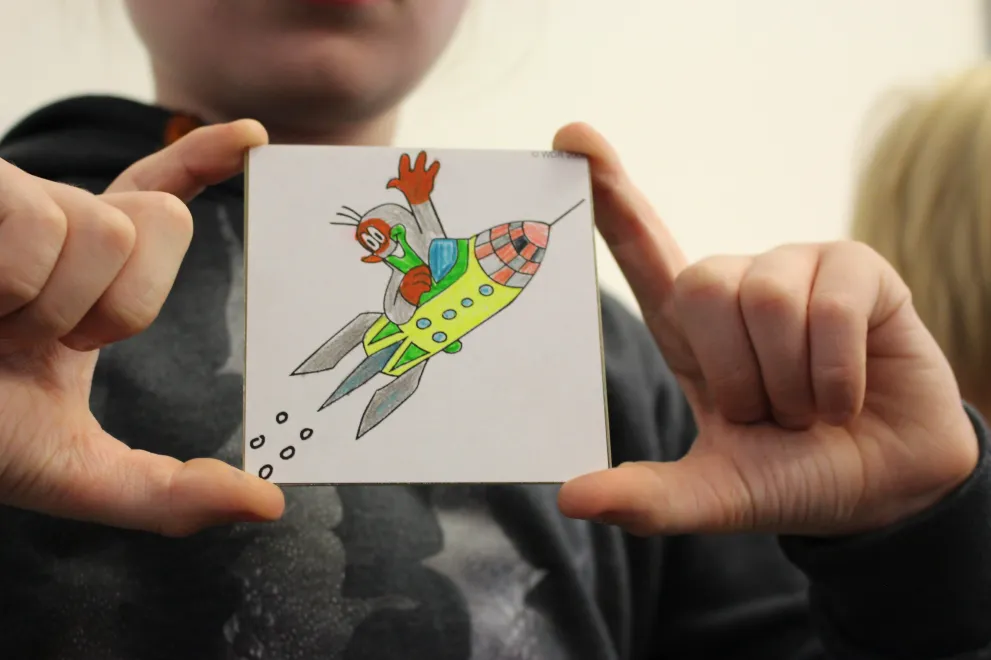Ein Kind zeigt ein selbstgemaltes Bild von einem Maulwurf auf einer Rakete.
