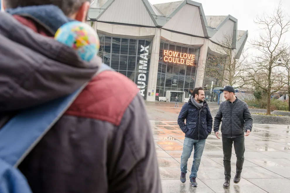 Weltkugel auf Schulter im Vordergrund, zwei Studierende unterhalten sich im Hintergrund. 