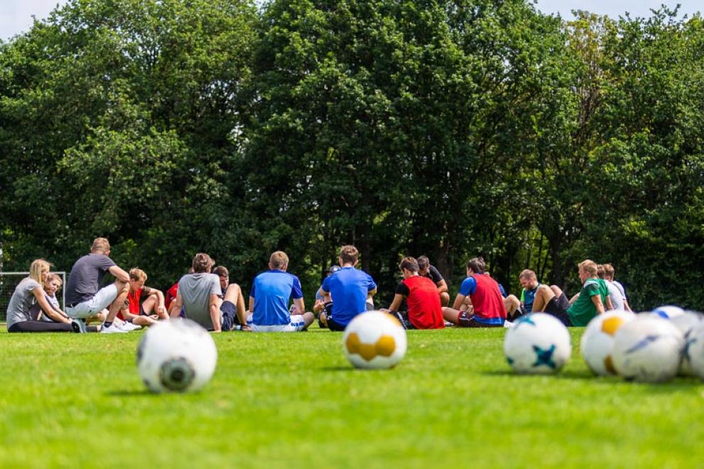 Sportstudierende sitzen im Kreis auf einer Wiese, im Vordergrund liegen Bälle
