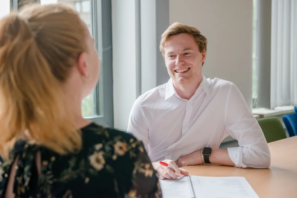 Ein junger Mann sitz am Tisch, vor sich Stift und Papier im Gespräch mit einer Frau, die man nur als Rückenfigur sieht.