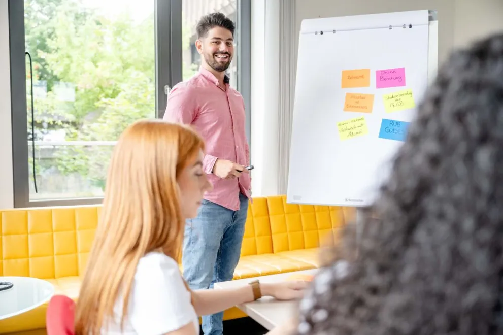 Junger Mann steht vor einer Gruppe Studierenden am Flipchart.