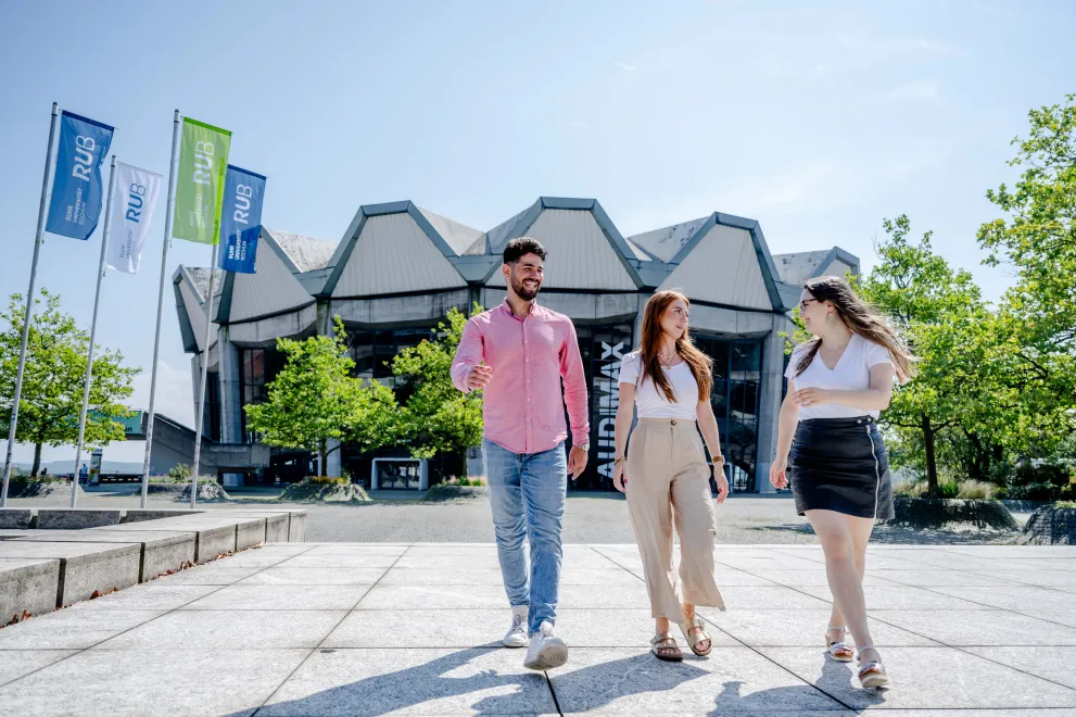 Drei Studierende laufen über den Forums-Platz vor dem Audimax und unterhalten sich.