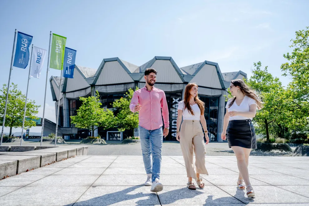 Three students walk across the forum square in front of the Audimax and talk.