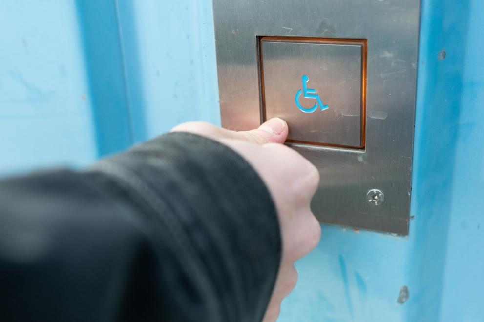 Foto: Druckknopf mit dem Symbol eines Rollstuhls in einem Fahrstuhl. Eine Hand drückt mit dem Daumen unten links auf den Druckknopf.
