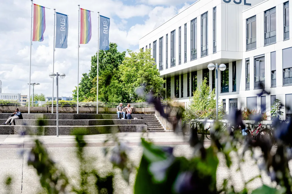Photo: Steps to sit on in front of the SSC, 3 people sitting on the steps. On the right, there is the Student Service Center (SSC). Above the steps, there are two flags with the RUB logo and two rainbow flags.