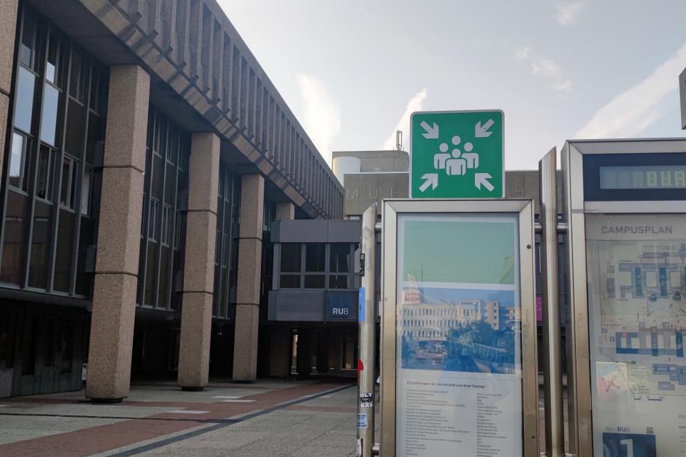 Signposting of an assembly point on the RUB campus. The icon shows four people in white on green, with four arrows pointing to them.