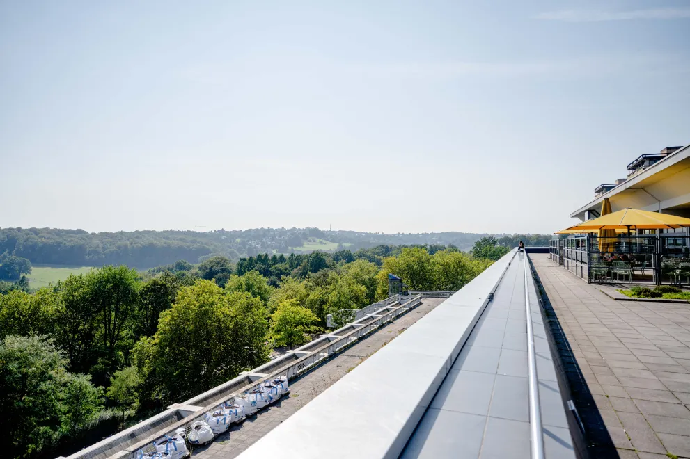 Photo: Terrace with seats, umbrellas and a railing, next to it a view of a valley with many green trees. It is sunny.