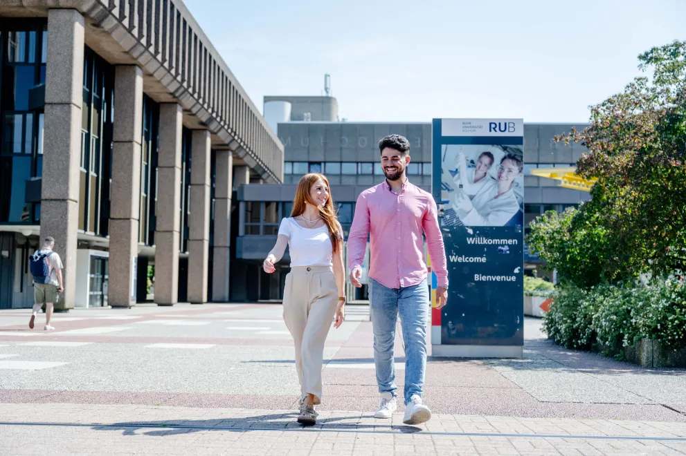Foto: Eine Frau und ein Mann gehen auf dem betonierten Campus der Ruhr-Universität nebeneinander her. Sie sind frontal zu sehen, sie unterhalten sich und lachen. Es ist sonnig. Im Hintergrund sieht man ein Schild mit der Aufschrift "Willkommen" in mehreren Sprachen.