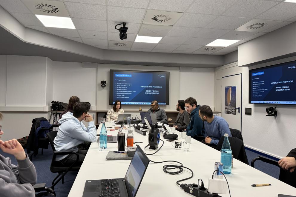 Teilnehmende des Workshops "Digitale Barrieren erkennen, benennen, vermeiden" sitzen gemeinsam mit den Referent*innen um einen großen Tisch, auf dem u.a. mehrere Laptops stehen, die an Mehrfachsteckdosen und einer Kabeltrommel angeschlossen sind.