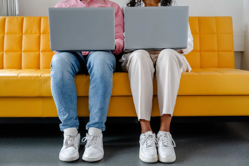 Foto: Zwei Personen sitzen nebeneinander. Beide haben einen aufgeklappten Laptop auf dem Schoß. Man sieht nur Füße und Beine der Personen sowie die Rückseite der Laptops.