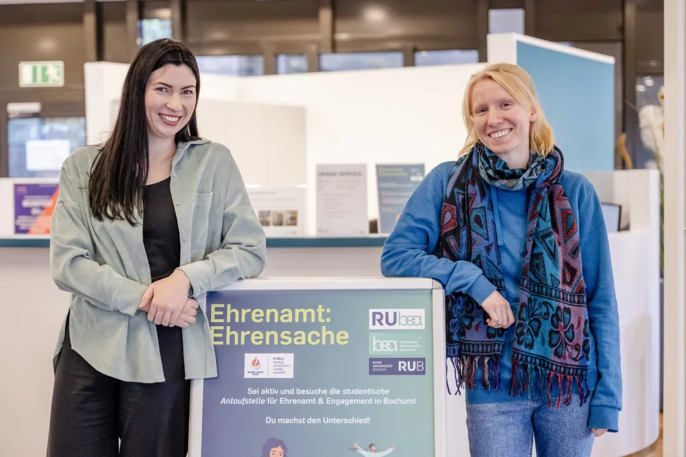 Zwei Studentinnen lehnen an einen Aufsteller und lächeln in die Kamera.