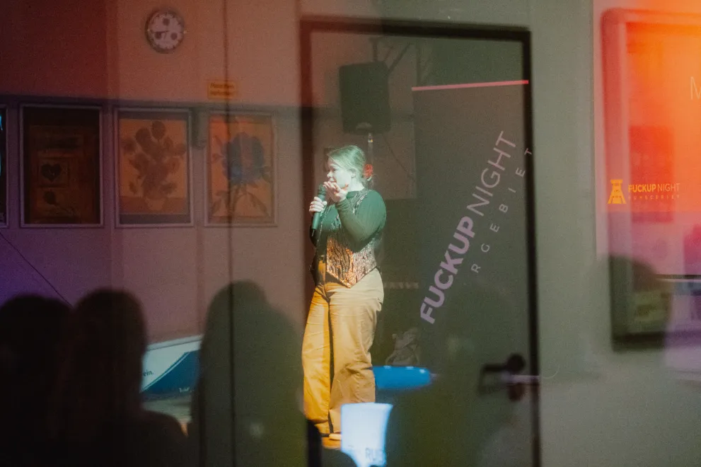 Foto durch eine Glasscheibe hindurch. Zu sehen ist eine Frau, die etwas auf einer Bühne erzählt.