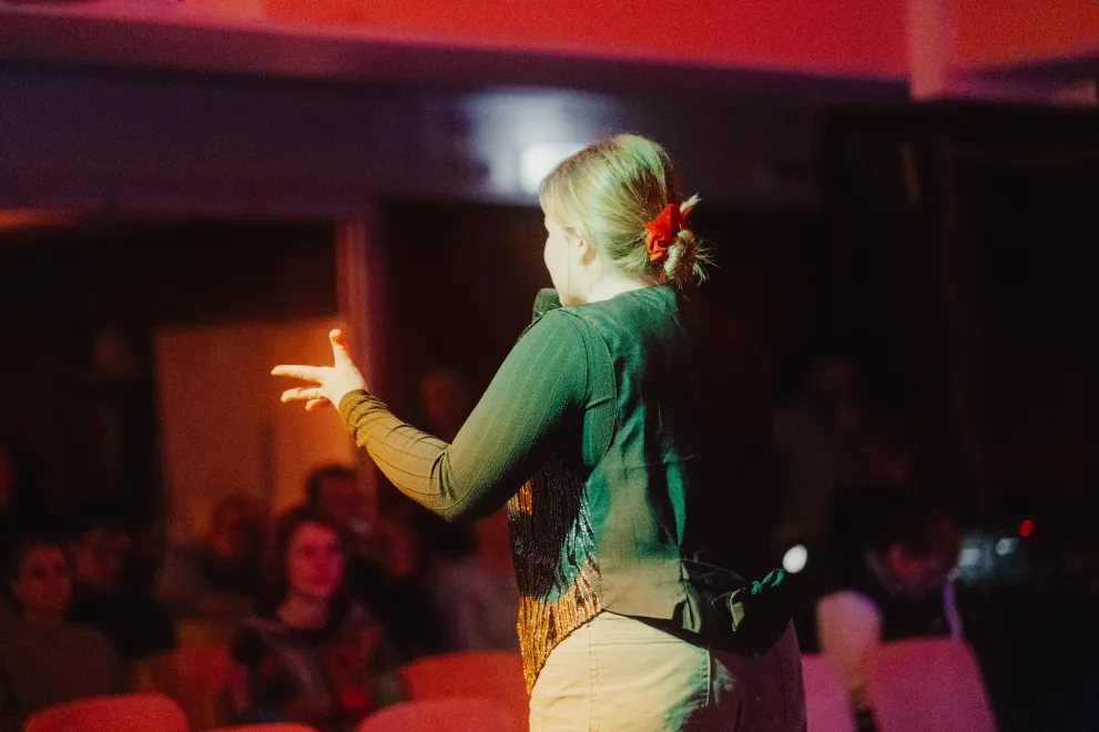 Rückansicht einer weiblich gelesenen Person, die auf einer Bühne steht und mit einem Mikrofon in der Hand Richtung Publikum spricht