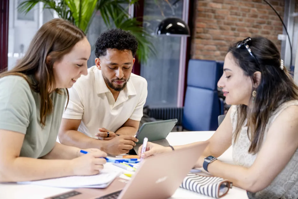Einer Gruppe von drei Studierenden sitzt mit Laptops und Unterlagen gemeinsam an einem Tisch.