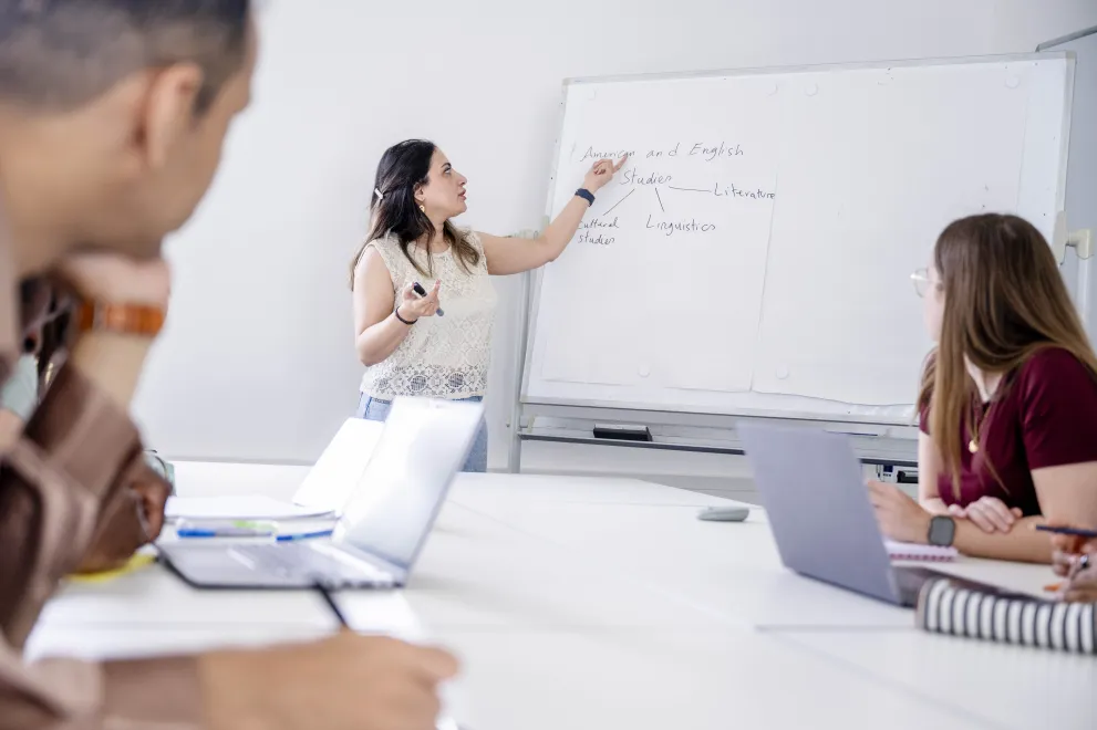 Eine Frau leitet eine Coaching-Sitzung an einem Flipchart vor einer Gruppe Studierender.