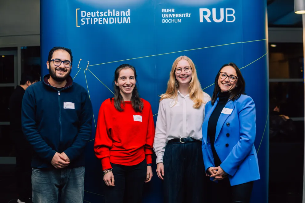 Foto: Vier Personen stehen vor einem blauen Aufsteller. Sie lächeln in die Kamera. Auf dem Aufsteller sind die Logos des Deutschlandstipendiums und der Ruhr-Universität Bochum zu sehen.