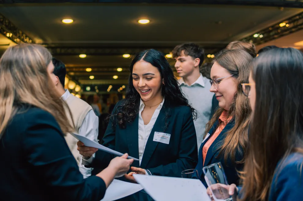 Foto: Eine Frau überreicht einer anderen Frau ein Dokument. Sie lächelt. Daneben stehen weitere Personen.