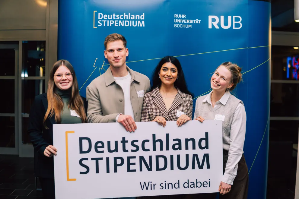 Foto: Vier Personen stehen vor einem blauen Aufsteller. Auf dem Aufsteller befinden sich das Logo des Deutschlandstipendiums und das Logo der Ruhr-Universität Bochum. Die vier Personen halten zusammen ein Schild mit der Aufschrift "Deutschlandstipendium - Wir sind dabei".