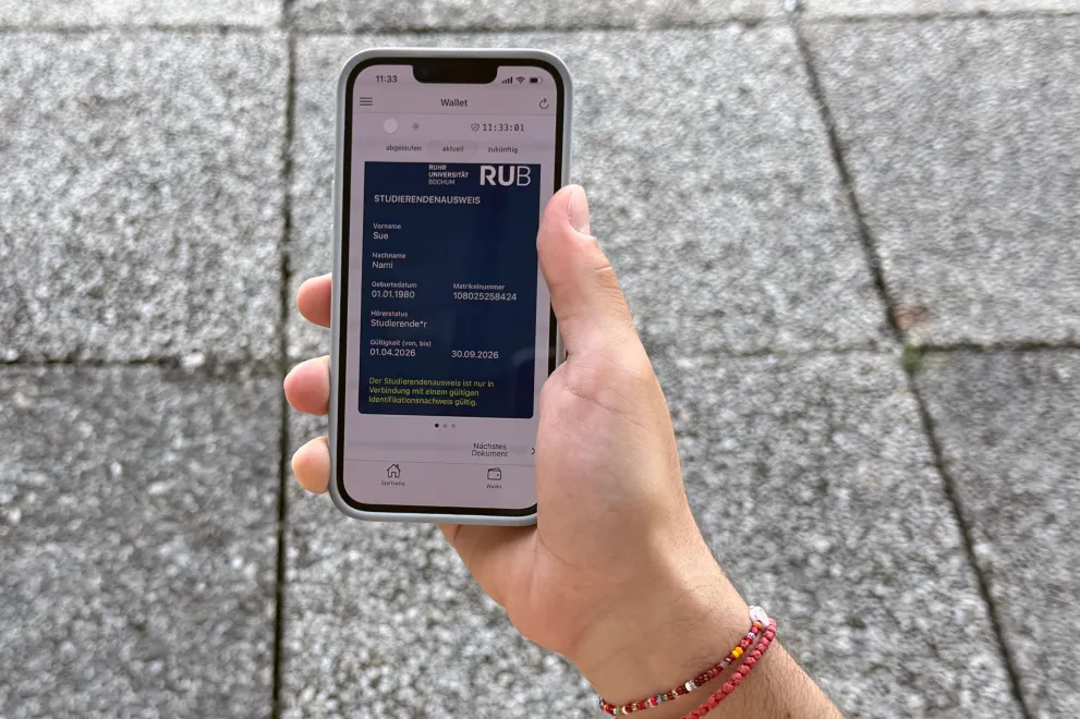 Das Bild zeigt eine Hand, die ein Handy hält, dahinter erkennt man die RUB-typischen Klapperplatten.  Auf dem Display des Handys ist ein  digitaler Studierendenausweis aufgerufen.