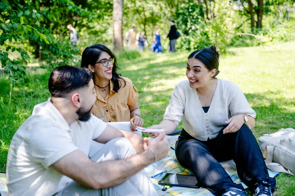 Das Foto zeigt zwei junge Frauen und einen jungen Mann die zusammen auf einer Decke auf einer Wiese sitzen und zusammen reden und lachen.