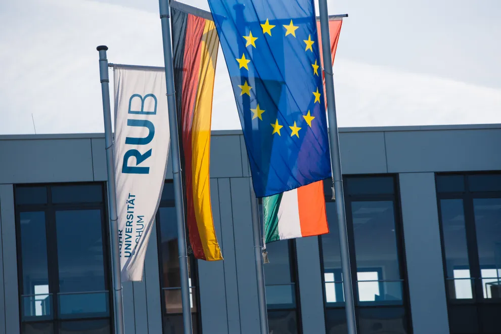 Das Foto zeigt verschiedenen Flaggen an Fahnenmasten vor einem Gebäude: Im Vordergrund die EU-Flagge, daneben Deutschland und RUB-Flagge, im Hintergrund verdeckt erahnt man noch die Landesflagge NRW
